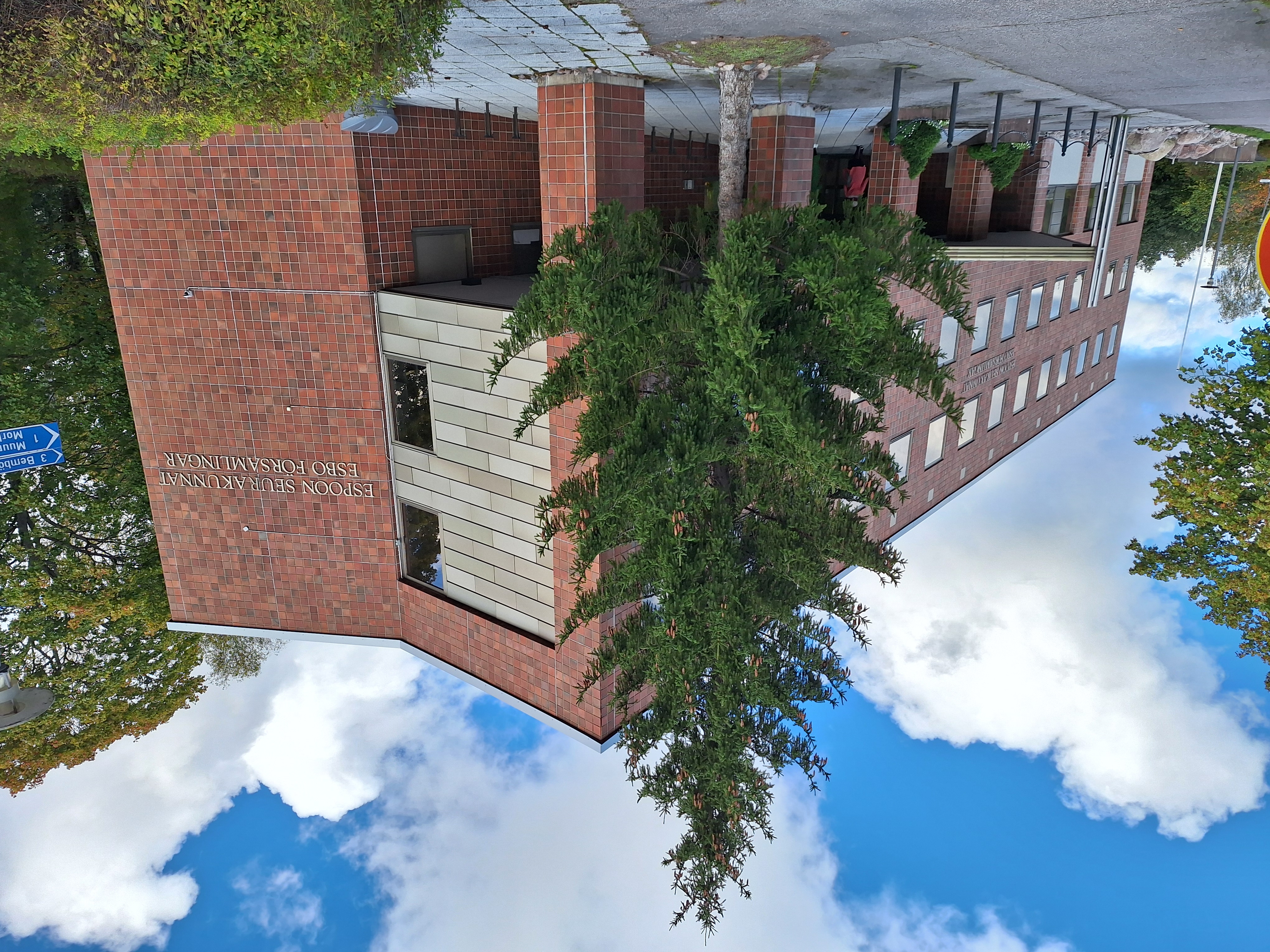En rödtegelbyggnad ed en stor gran framför och blå himmel.