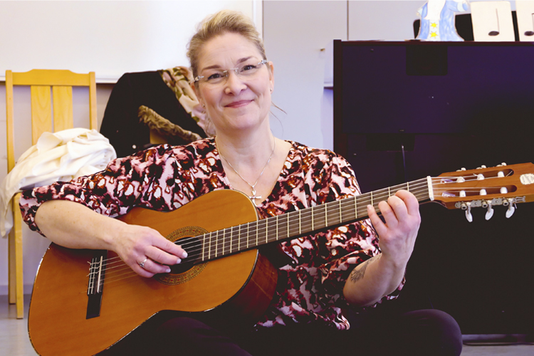 En kvinna med blont hår och glasögon sitter på golvet med en gitarr i famnen.