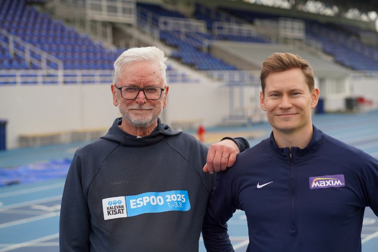 Två män står i mörka tröjor på en arena med läktare i bakgrunden.