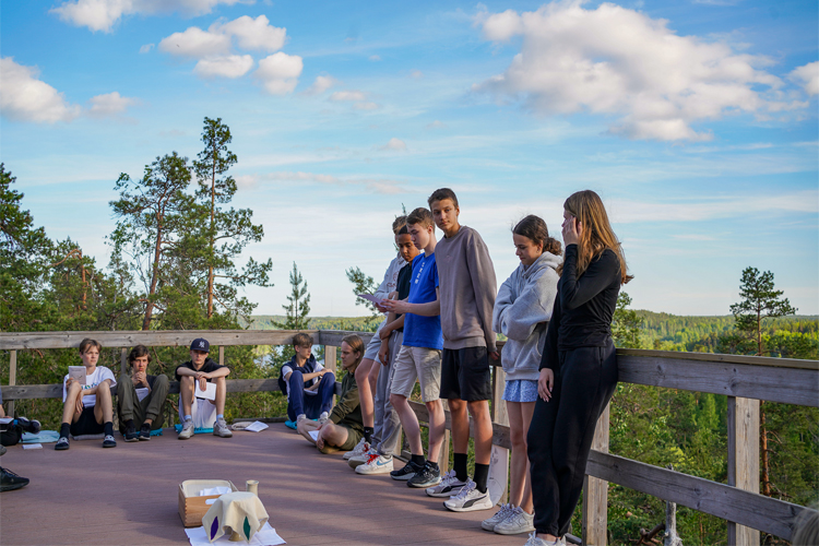 Unga står och sitter på en terass utomhus som är i höjd med trädtopparna och blå himmel bakom.