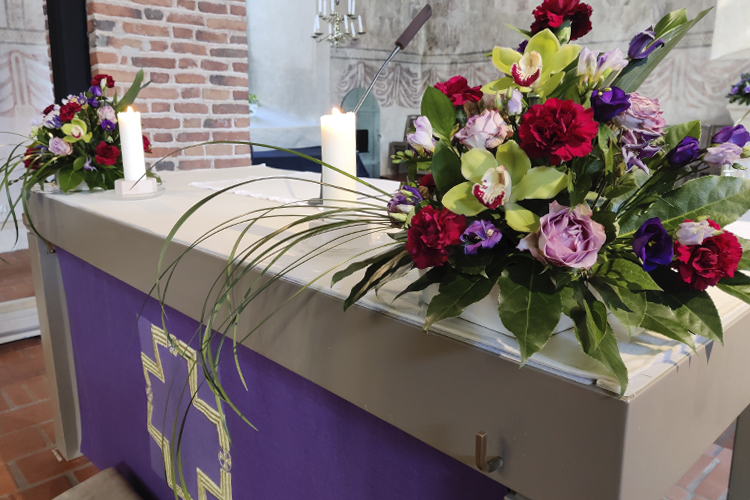 Närbild av altaret med lila duk, blomsterarrangemang och två tända ljus.