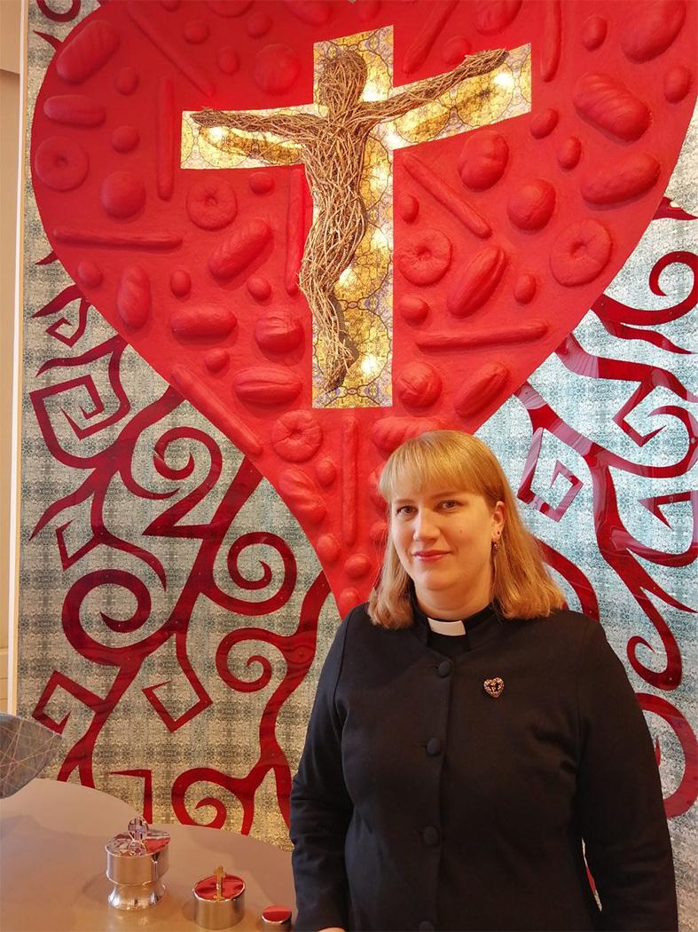 Kira Ertman står intill altaruppsatsen, ett rött hjärta med krucifix i guld i Karabacka kapell.
