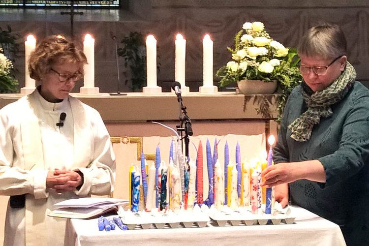 Handstöpta ljus i olika färger tänds på altaret. Två kvinnor varav den ena är präst.