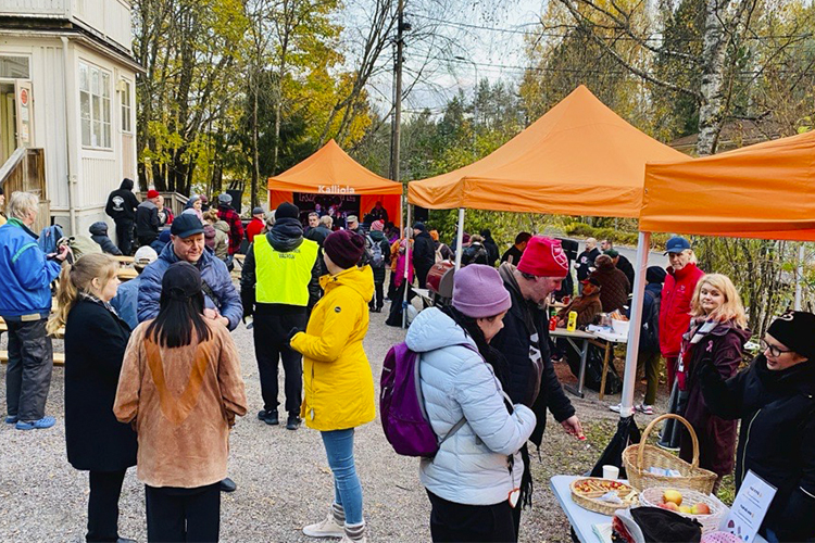 Tiotals människor står på en gårdsplan utanför en trävilla och på gården orangefärgade tält.