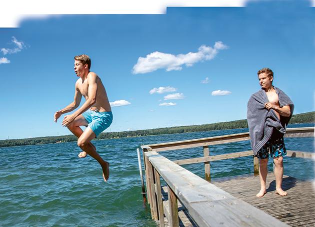 Två pojkar i badbyxor. Den ena står på bryggan medan den andra hoppar i sjön.