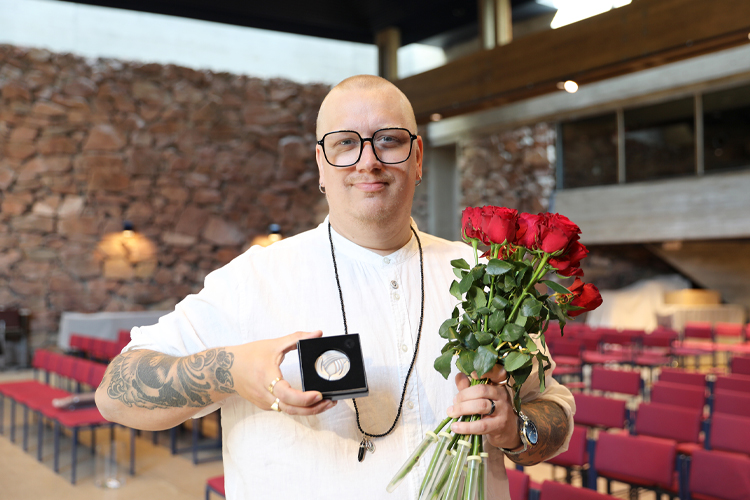 Arttu Wiskari iklädd vit tskjorta och glasögon håller upp medaljen i sin ask framför sig och håller en buke...