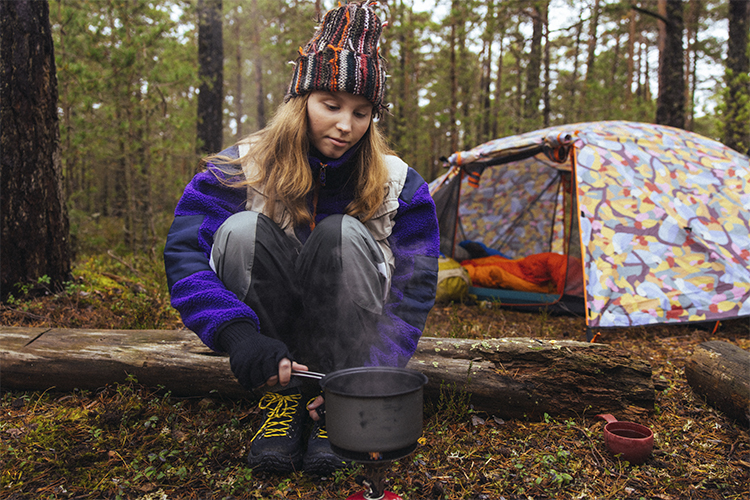 En ung flicka sitter friluftsklädd i skogen, med en kastrull i handen på huk intill ett tält.