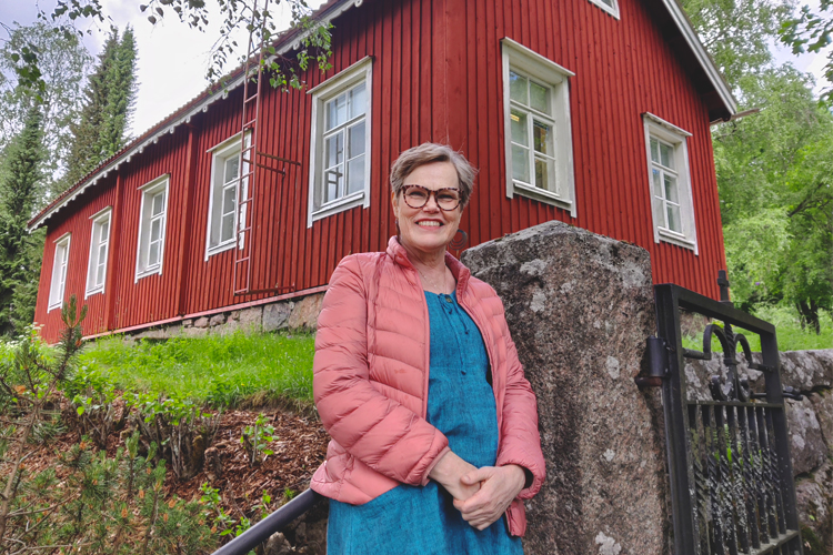 En kvinna i rosa täckjacka lutar sig mot en räcke framför en rödmålad träbyggnad i allmogestil intill Esbo ...