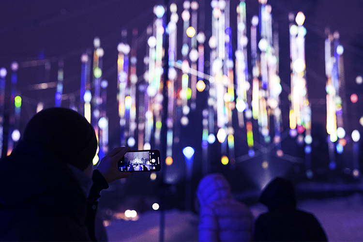 Lodräta ljusprismor i lilafärgat mörker fotograferas av personer med smarttelefon.
