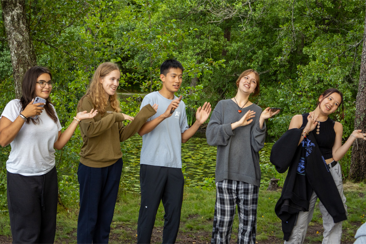 Fem leende unga står utomhus i grönska och gör en vågrörelse med händerna.