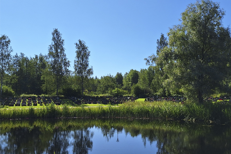 Klockarmalmens gravplatser syns på avstånd i sommargrönska och i förgrunden en spegelblank sjö.
