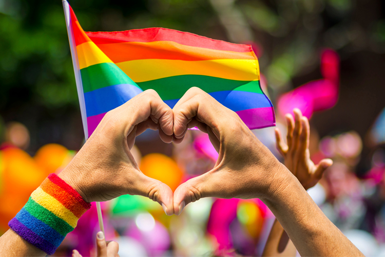 En Prideflagga vajar ovanför folkhav och någon gör hjärtsymbolen med sina händer framför flaggan.