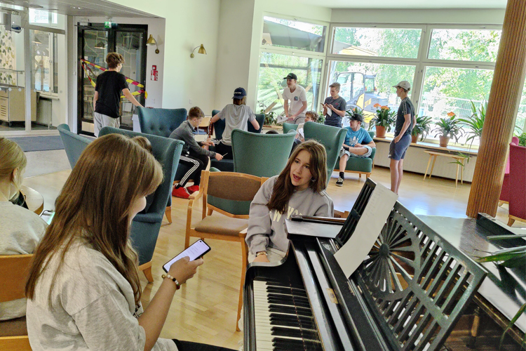 Unga sitter inomhus och umgås, en flicka sitter vid ett piano och tittar på sin mobil.