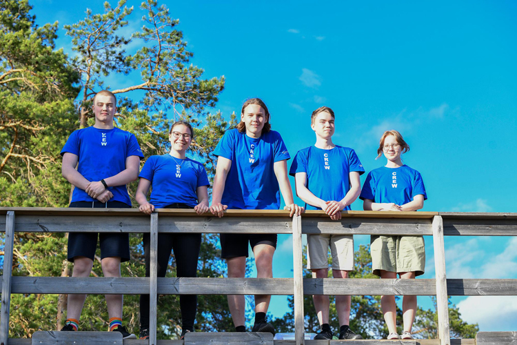 Fem unga står i blås t-skjortor på en terrass, mot blå himmel i bakgrunden och tallkvistar.