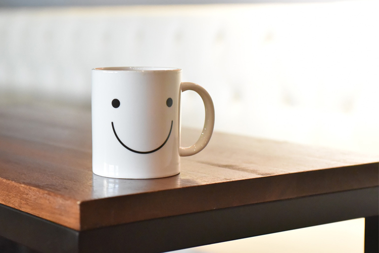 En vit mugg med en smiley-emoji på står på en bordsskiva i mörkbrunt trä.