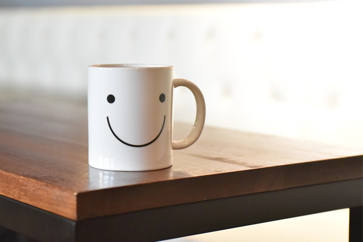 En vit porslinsmugg med en smile-emoji står på ett brunt träbord.