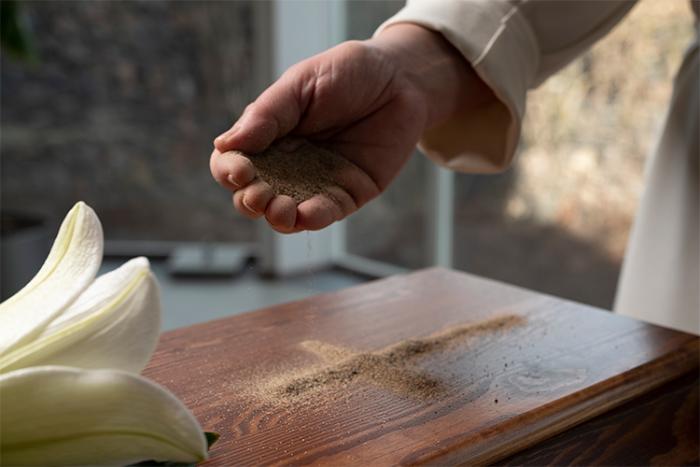 Prästens hand tecknar ett kors av sand på en kista med en vit lilja på locket.