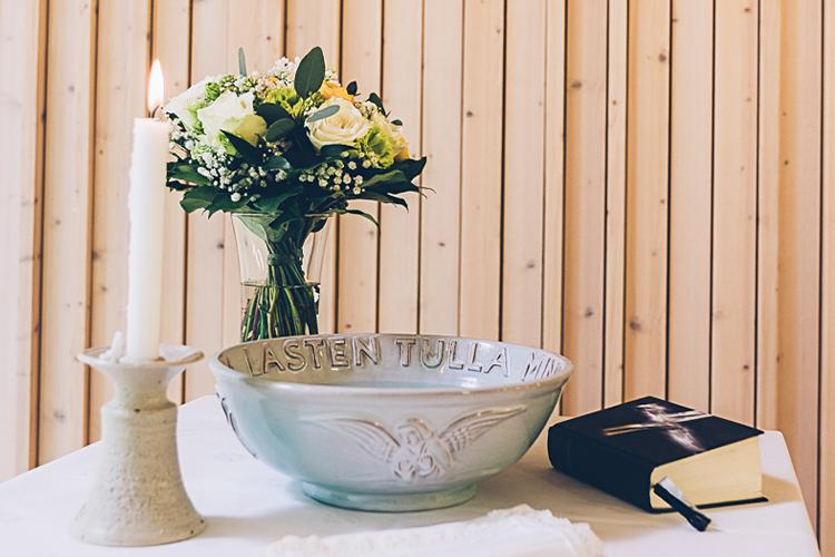 En dopskål på ett bord med vit duk, en ljusstake med ett tänt ljus, en blombukett och en bibel.