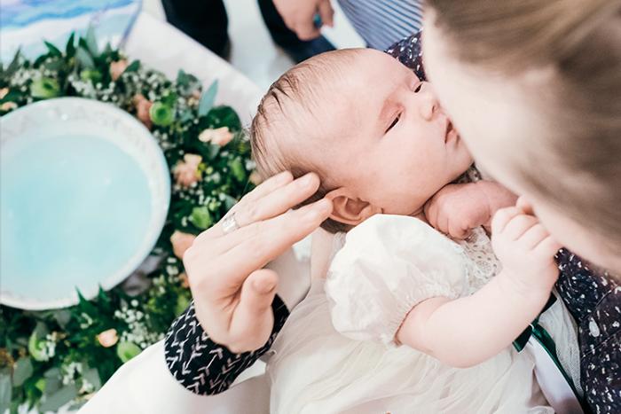 Liten baby i dopklänning i en vuxen persons famn. En dopskål prydd med en blomkrans finns med på bilden.