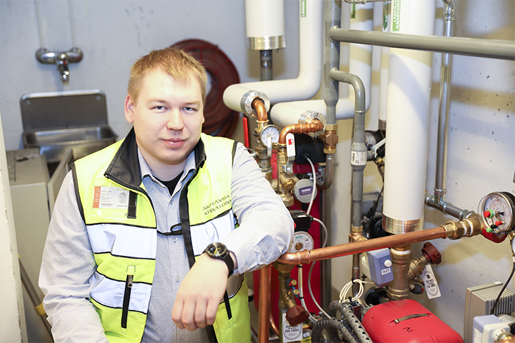 Harri Heiskanen sitter i arbetskläder i en värmecentral intill rör och mätare.