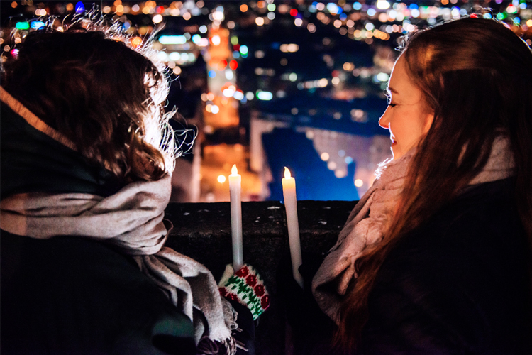 Två flickor fotade bakifrån som sitter med ljus i händerna och blickar ut över en stadsvy med ljuspunkter i...