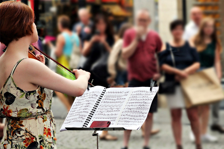 Ung kvinna spelar violin med ett notställ framför sig och människor som promenerar förbi på gatan.