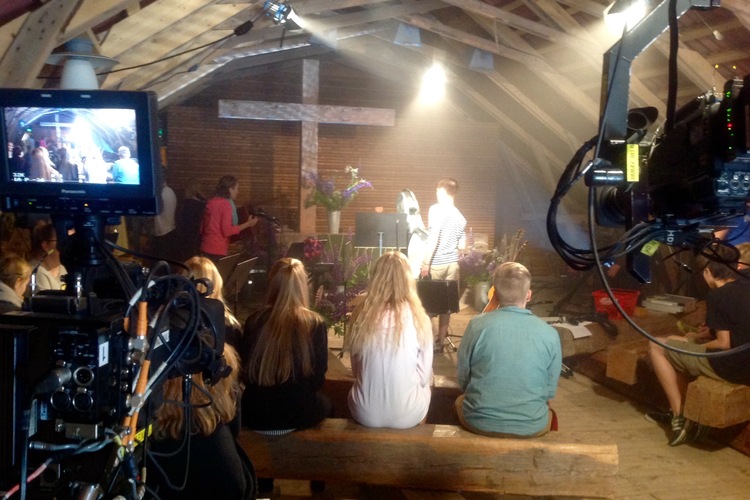 Ett litet gudstjänstutrymme där människor sitter på golvet. En tv-skärm och ett kors skymtar i bakgrunden.