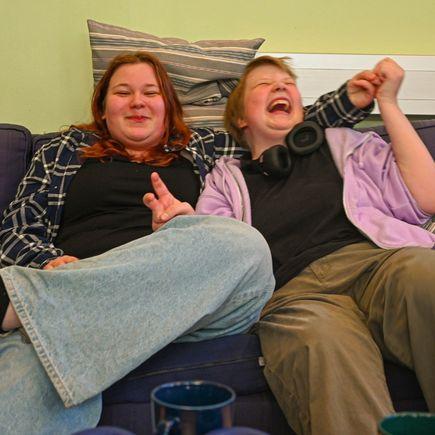 Två unga personer sitter på en soffa. Den ena ler och den andra skrattar hejdlöst.